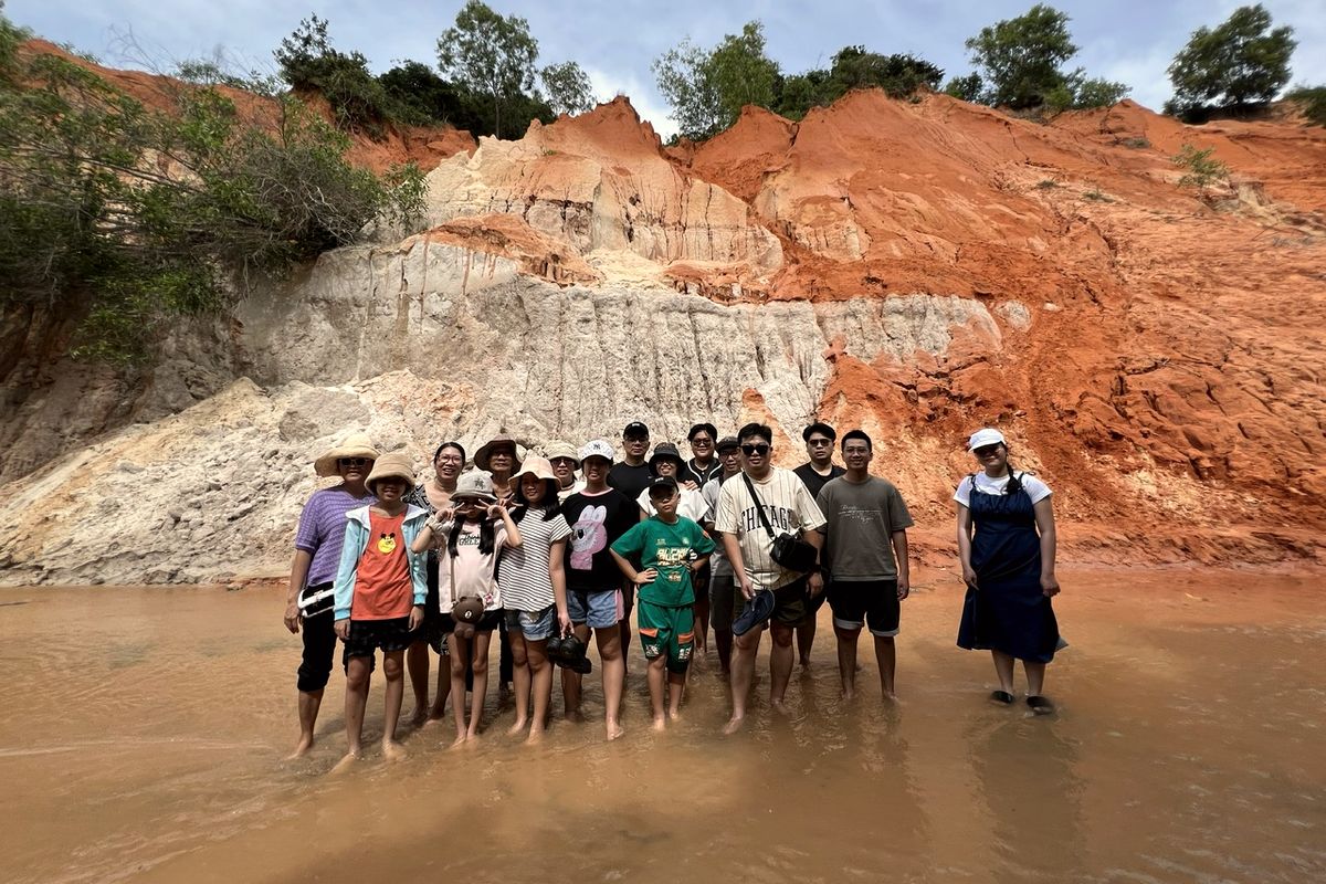 Đoàn Sài Gòn Tâm Điểm và người thân đứng chân trần giữa dòng suối Tiên, phía sau là vách cát trắng và đỏ