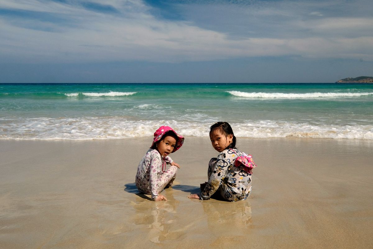 Hai bé gái ngồi chơi cát bên mép nước biển xanh ở Cam Ranh